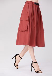 Jupe midi rouge en tissu léger, dotée d'une taille haute, de poches latérales et d'une ceinture. Associée à des escarpins noirs à lanières.