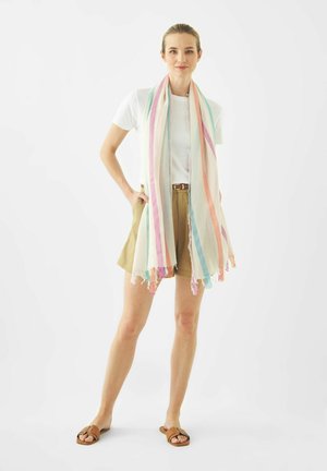 Vrouw draagt een wit T-shirt, beige short, bruine sandalen en een lange sjaal met pastelstrepen, staand tegen een effen witte achtergrond.