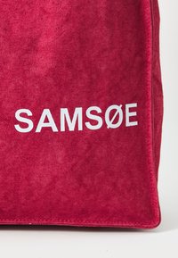 Borsa in tessuto rosso con superficie testurizzata, caratterizzata dal nome del marchio "SAMSOE" in lettere bianche in grassetto su un lato.