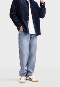 Man in een marineblauw corduroy shirt, witte T-shirt, losse lichtblauwe jeans en witte sneakers met zwarte strepen, staand met één hand in zijn zak.