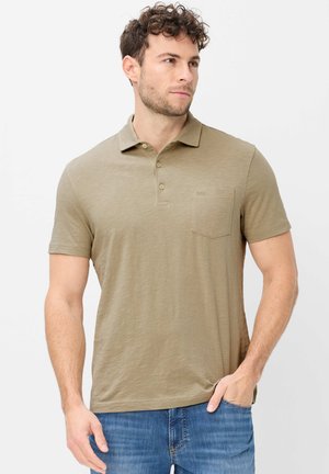 Mand iført lysebrun kortærmet poloshirt med brystlomme, stående med venstre hånd i jeanslomme, kiggende til siden.