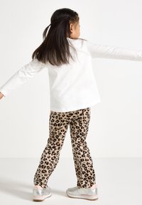 Hvit langermet skjorte og beige leopardmønstret bukser på et barn. Sølvfargede sneakers med rosa detaljer. Bakfra, med armene utstrakt.