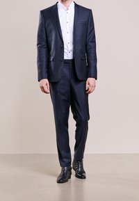 Completo blu navy con un sottile motivo a pois, giacca a un bottone e pantaloni slim-fit. Abbinato a una camicia bianca e scarpe nere.