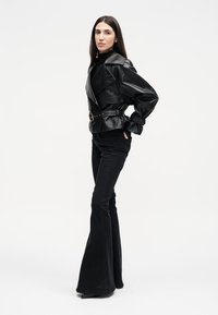 Γυναίκα όρθια με τα χέρια στις τσέπες, φορώντας μαύρο δερμάτινο μπουφάν με ζώνη, μαύρο ζιβάγκο και μαύρο φαρδύ παντελόνι, σε λευκό φόντο.