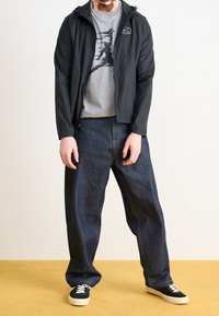 Černá lehká bunda s kapucí, šedé tričko s grafikou, volné modré džínové kalhoty a černé tenisky s bílými podrážkami na žluté podlaze.