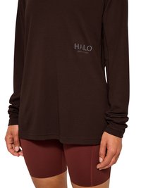 Langärmliges schwarzes, strukturiertes Oberteil mit „HALO“-Logo kombiniert mit bordeauxfarbenen Biker-Shorts bei einer stehenden Person, Hände entspannt an den Seiten.