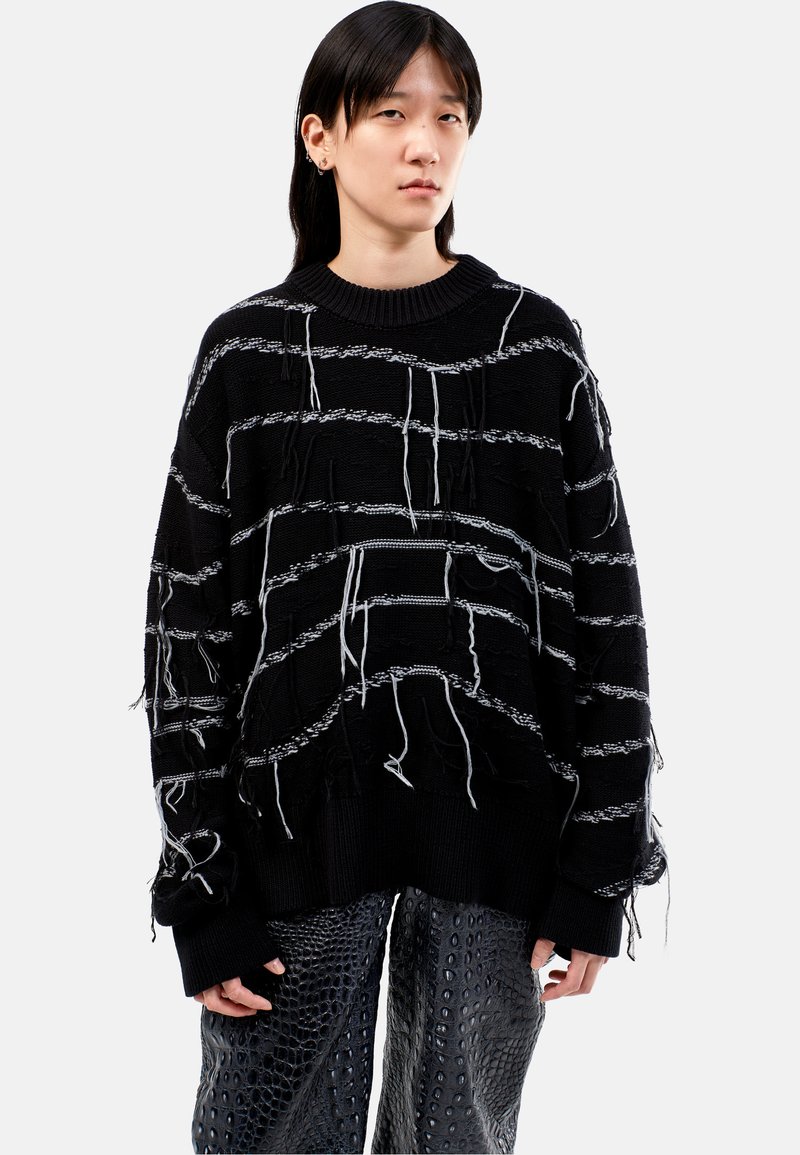 Zwarte gebreide oversized trui met witte horizontale strepen en hangende draadjes. Gepaard met gestructureerde zwarte broek met een reptielpatroon.