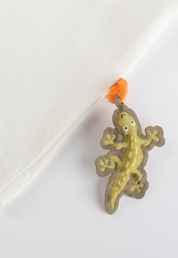 Cremfarbener Stoff mit einem gewebten Gecko-förmigen Schmuck in Grün und Grau, der oben mit einem orangenen Pompon versehen ist und nahe dem Rand befestigt ist.