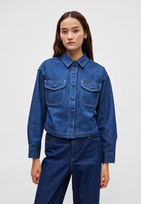 Sötétkék farmerdzseki, gallérral, gombos elülső résszel, két mellzsebbel és hosszú ujjakkal, amelyek manszettel végződnek.