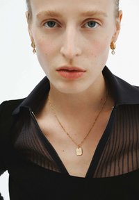 Primer plano de una persona que lleva pendientes de aro retorcidos de oro y un collar de oro con un colgante rectangular, vestida con una blusa negra con un cuello transparente.