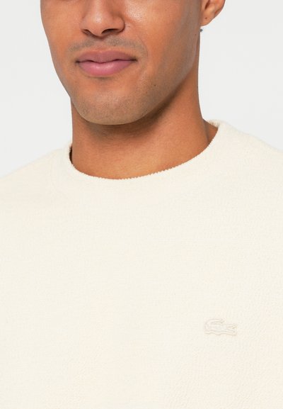 Sweatshirt en polaire couleur crème avec un col rond et une surface texturée. Présente un logo brodé subtil sur la poitrine.