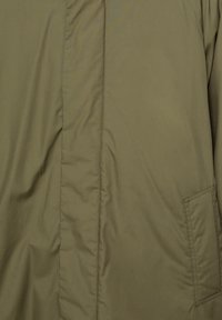 Giacca imbottita verde oliva con una superficie liscia, tasche laterali e dettagli di cucitura visibili sul pannello frontale. Il tessuto è leggero e resistente.