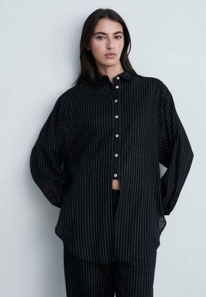 Chemise noire à boutons avec des rayures blanches verticales, coupe loose, manches longues et col, présentée sur un fond neutre.