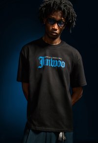 Černé bavlněné tričko s modrým grafickým textem „Solo Leveling Jinwoo“. Model má tmavé sluneční brýle; další detaily zahrnují zipy a řetízkové ozdoby.