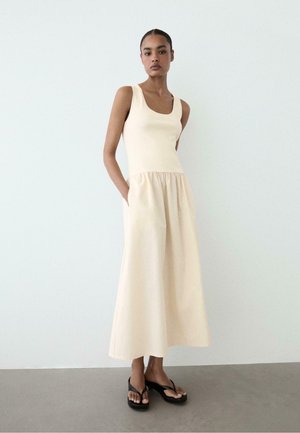 Frau trägt ein ärmelloses cremefarbenes Maxikleid mit Taschen, steht vor einer schlichten hellen Wand und trägt schwarze Plateausandalen.