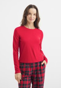 Red long-sleeve top with a ribbed texture, featuring a round neckline and a slightly cropped hem, paired with plaid pants in red, green, and black.