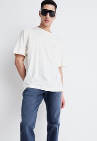 Camiseta de algodón crema de manga corta, con un gráfico sutil. Combinada con jeans de mezclilla azul oscuro. El modelo lleva puestas unas gafas de sol negras y de gran tamaño.