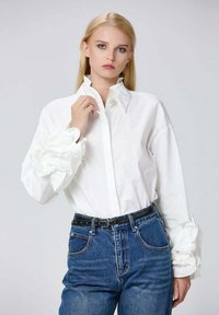 Chemise en coton blanche avec col haut et poignets froncés à motifs, assortie à un jean en denim bleu taille haute et une ceinture noire.