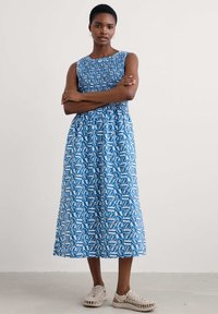Maxi dress in blue with white leaf patterns, featuring a smocked bodice and sleeveless design. Paired with light woven shoes.