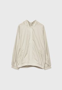 Veste beige clair zippée avec poignets élastiques, deux poches avant et un bas légèrement froncé, présentée sur un fond uni.