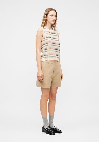 Γυναίκα με κυματιστά ξανθά μαλλιά, που φοράει αμάνικο ριγε πλεκτό τοπ, μπεζ σορτς μέχρι το γόνατο, γκρι κάλτσες και μαύρα μοκασίνια, στέκεται πλευρικά.