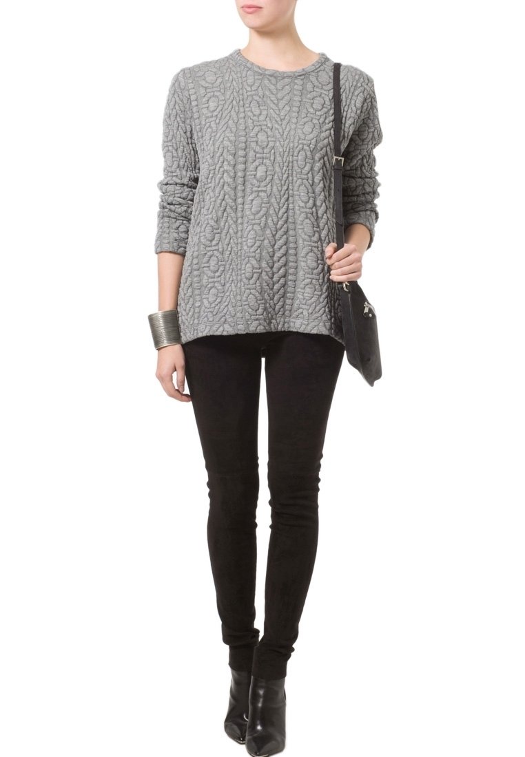 Femme portant un pull gris en maille torsadée, un pantalon slim noir, des bottines noires, un bracelet manchette argenté et un sac à bandoulière noir.