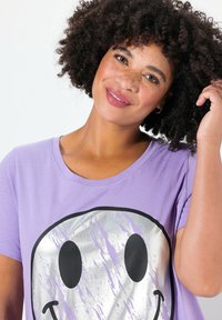 T-shirt en coton violet avec un grand motif de smiley argenté agrémenté d'accents noirs. Texture douce et coupe décontractée.
