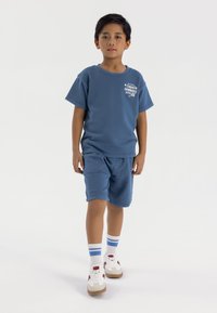 Maglietta di cotone blu con stampa grafica, abbinata a pantaloni corti blu coordinati, sneakers bianche e rosse, e calze a righe fino alla caviglia.