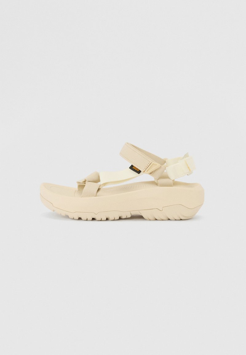 Beige utendørs sandal med tykk gummisåle, justerbare stropper og en teksturert overflate. Designet legger vekt på komfort og stabilitet.
