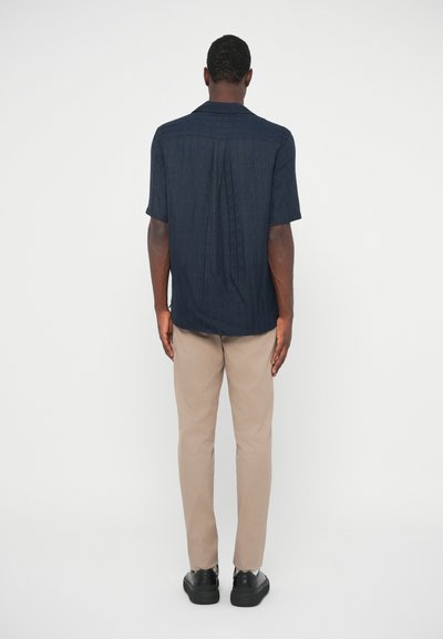 Kortärmad mörkblå skjorta med en subtil textur, med rak fåll och bakveck, i kombination med ljusbeige byxor och svarta skor.