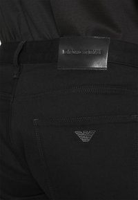 Black denim pants with a smooth texture, featuring a leather label at the waist and a subtle, embossed logo on the back pocket.