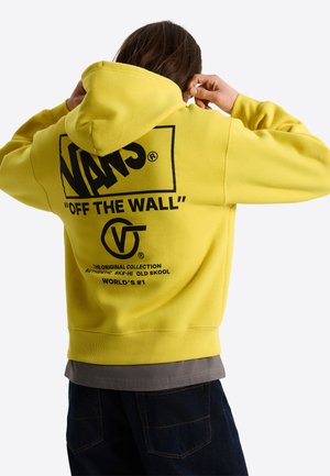 Personne portant un sweat à capuche Vans jaune vif avec texte et logos noirs dans le dos, ajustant la capuche, de dos à la caméra.