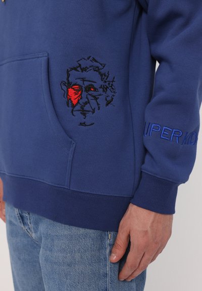 Felpa blu con tasca frontale, caratterizzata da un volto ricamato in rosso e nero. Include la scritta "SUPERM" ricamata in blu sulla manica.