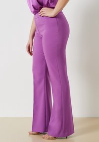 Lila flarebyxor med ett mjukt, stretchigt material. Hög midja. Synlig sömnad längs sömmarna, tillsammans med snörda sandaler.