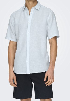 Homme portant une chemise bleu clair à manches courtes avec boutons et un short noir arrivant aux genoux, debout devant un fond blanc uni.