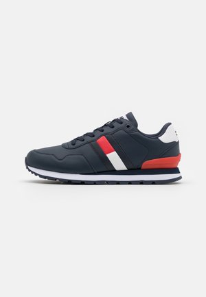 Zapatilla deportiva azul marino con cordones negros, rayas laterales blancas y rojas, lengüeta blanca en el talón y detalles en rojo y azul marino en la suela.