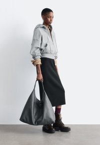 Grauer Zip-Hoodie mit übergroßen Ärmeln, schwarzer Midirock, dunkelbraune Schnürstiefel und eine große graue Ledertasche.