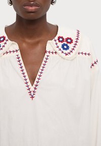 Primer plano de una persona que lleva una blusa blanca con escote en V y patrones florales bordados en rojo y azul en el cuello y las costuras.