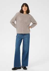 Maglione beige lavorato a maglia con motivo a rombi, colletto rotondo e polsini a costine, abbinato a jeans in denim blu a gamba larga e scarpe nere.