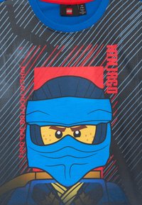 Lego Ninjago varonis ar zilu ninjas masku un intensīvām acīm, uz melna krekla ar diagonālām svītrām.