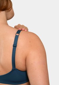 Soutien-gorge teal avec des bretelles réglables vu de dos ; texture lisse, fermeture à crochets et un détail en anneau rond près de la bretelle.