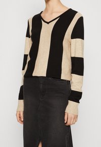 Sweter w paseczki w odcieniach beżu i czerni, z dekoltem w serek, długimi rękawami i ściągaczami przy mankietach. Noszony na czarnej spódnicy z denimu.