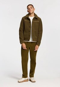 Jachetă din fleece verde măsliniu cu închidere cu nasturi și guler în picioare, asortată cu pantaloni din catifea cu caneluri și adidași bej. Textură netedă.