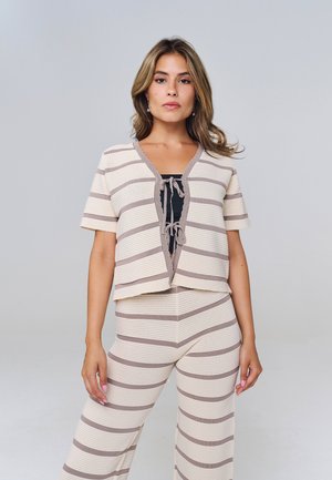 Vrouw staand, draagt beige en taupekleurig horizontaal gestreept gebreid kort mouwen top en bijpassende wijde broek, met zwart topje eronder, grijze achtergrond.