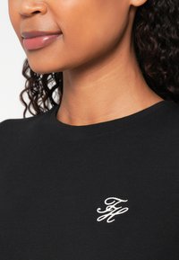 Tricou negru din bumbac cu guler rotund, având un mic logo brodat alb pe piept. Textură fină și croială ajustată.
