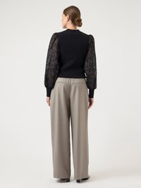 Woman with hair in bun wearing black fitted top with sheer floral puff sleeves and beige wide-leg pants, standing facing away on white background.