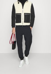 Veste noire et crème doublée en polaire avec fermeture éclair et deux poches zippées, associée à un jogging foncé et des baskets montantes blanches. Sac rouge.