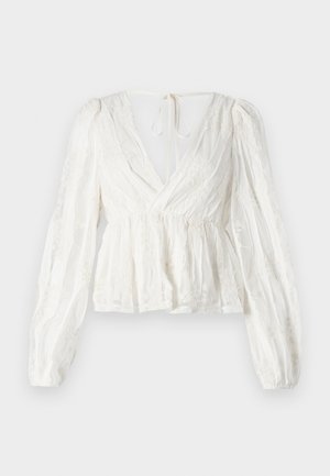 White blouse with a deep V-neck, long puffed sleeves, elastic waistband, and floral embroidery details. Features a tie closure at the back.