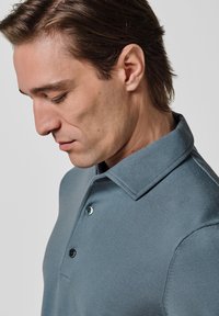 Poloshirt aus weichem, strukturiertem, gedämpftem blauem Stoff. Kurzer Kragen mit drei schwarzen Knöpfen. Nahaufnahme, die sich auf die Details des Hemdes konzentriert.
