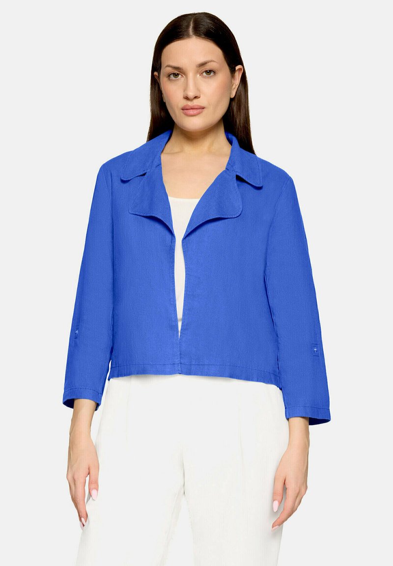 Femme aux cheveux bruns lisses portant une veste bleu vif ouverte sur un haut blanc et un pantalon blanc, debout devant un fond uni.